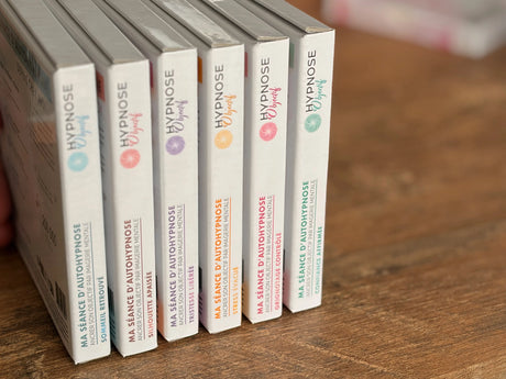 Collection de box HypnoseObjectif alignées sur une table en bois, représentant les six séances d’auto-hypnose : sommeil, stress, grignotage, tristesse, silhouette et confiance.